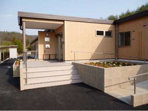 ベージュの壁に濃い茶色の屋根が載った平屋の建物で、玄関までは手すり付きの階段か、花壇を回り込むように設置されたスロープを利用する。