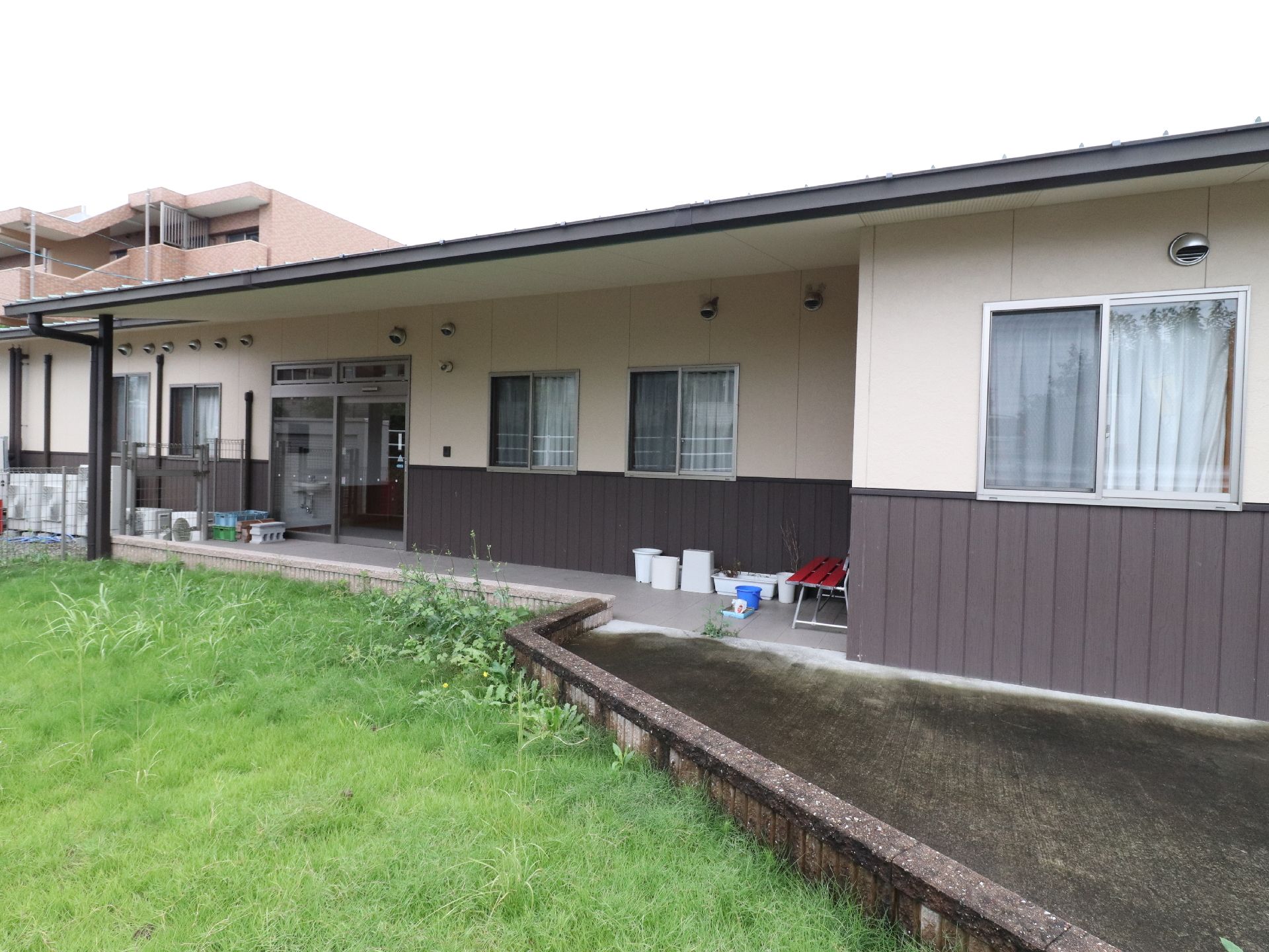 茶とクリームを基調とした平屋の施設の外観。玄関前には緑が広がっており、アットホームな雰囲気である。赤のベンチが置かれている。
