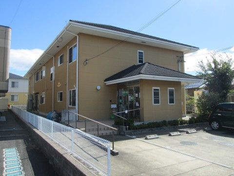 ベージュの外壁に切妻屋根がのった２階建の建物で、玄関部分が外に飛び出している。玄関前までは手すり付きのスロープか階段を利用する。