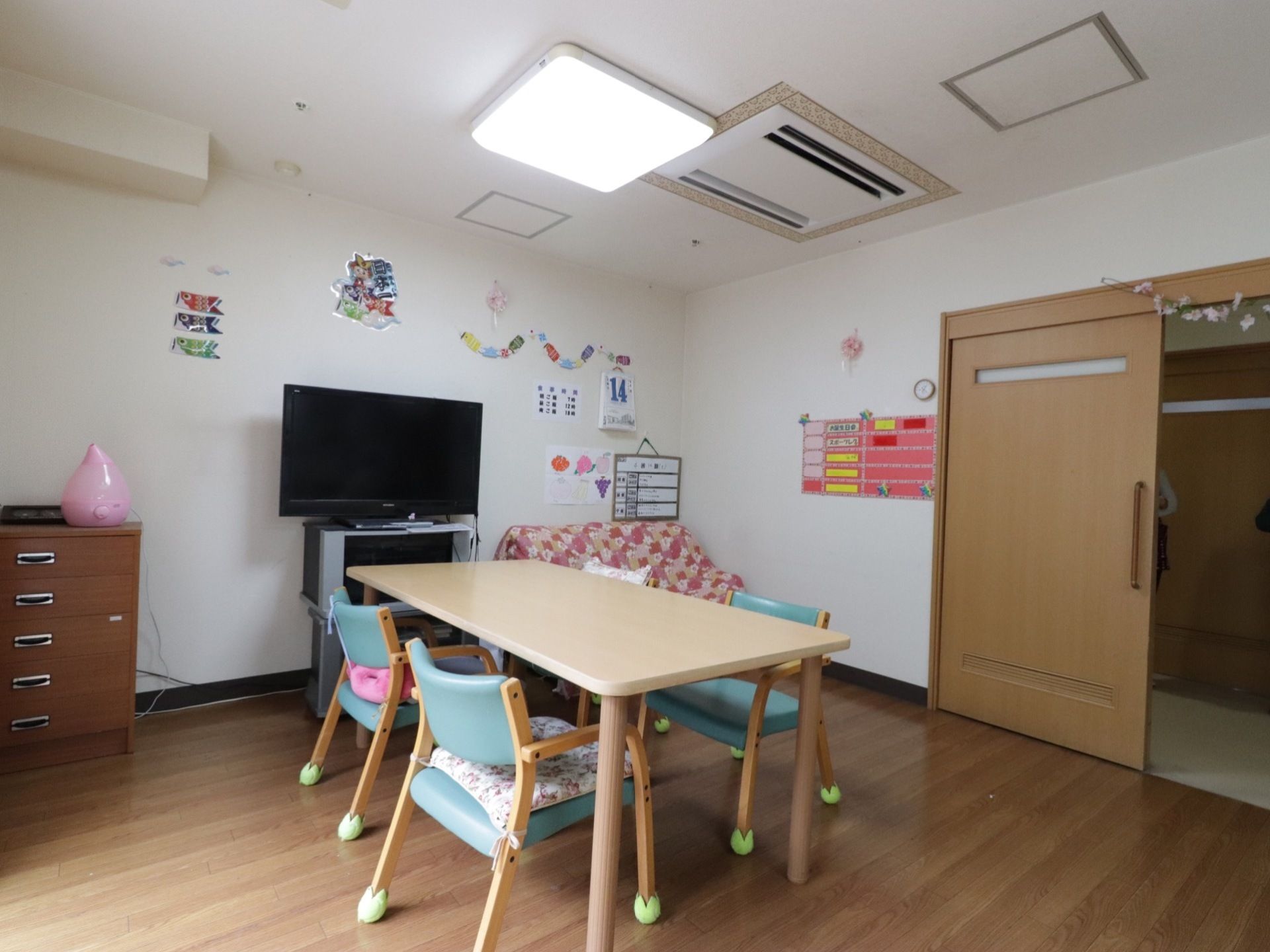 テレビの前には、テーブルとクッション材をつけた椅子がセットしてある。壁には掲示物があり入居者様が交流を持てる場所である。