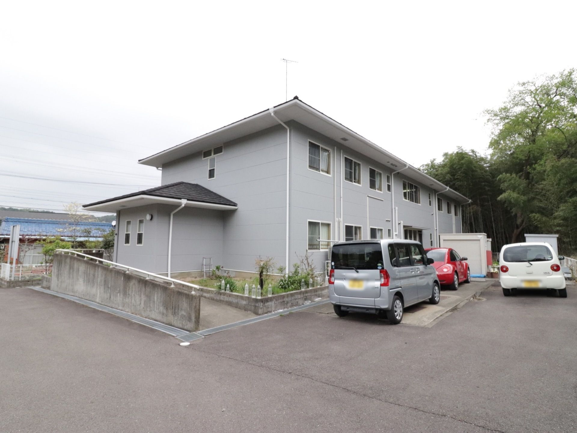 ２階に屋根があり、グレーの外壁に白い窓枠のシンプルな設計を採用している。駐車場からはスロープが伸びている。