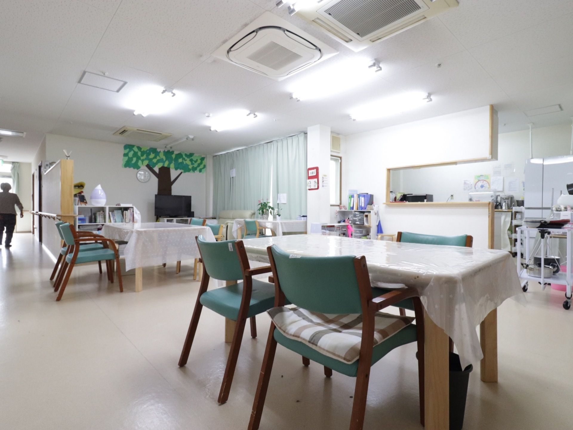 広い部屋に大きなテーブルが２つ、窓際に細長いテーブルが１つある。台所と併設してあるのでその壁の一部が空いている。窓際の近くにテレビがある。