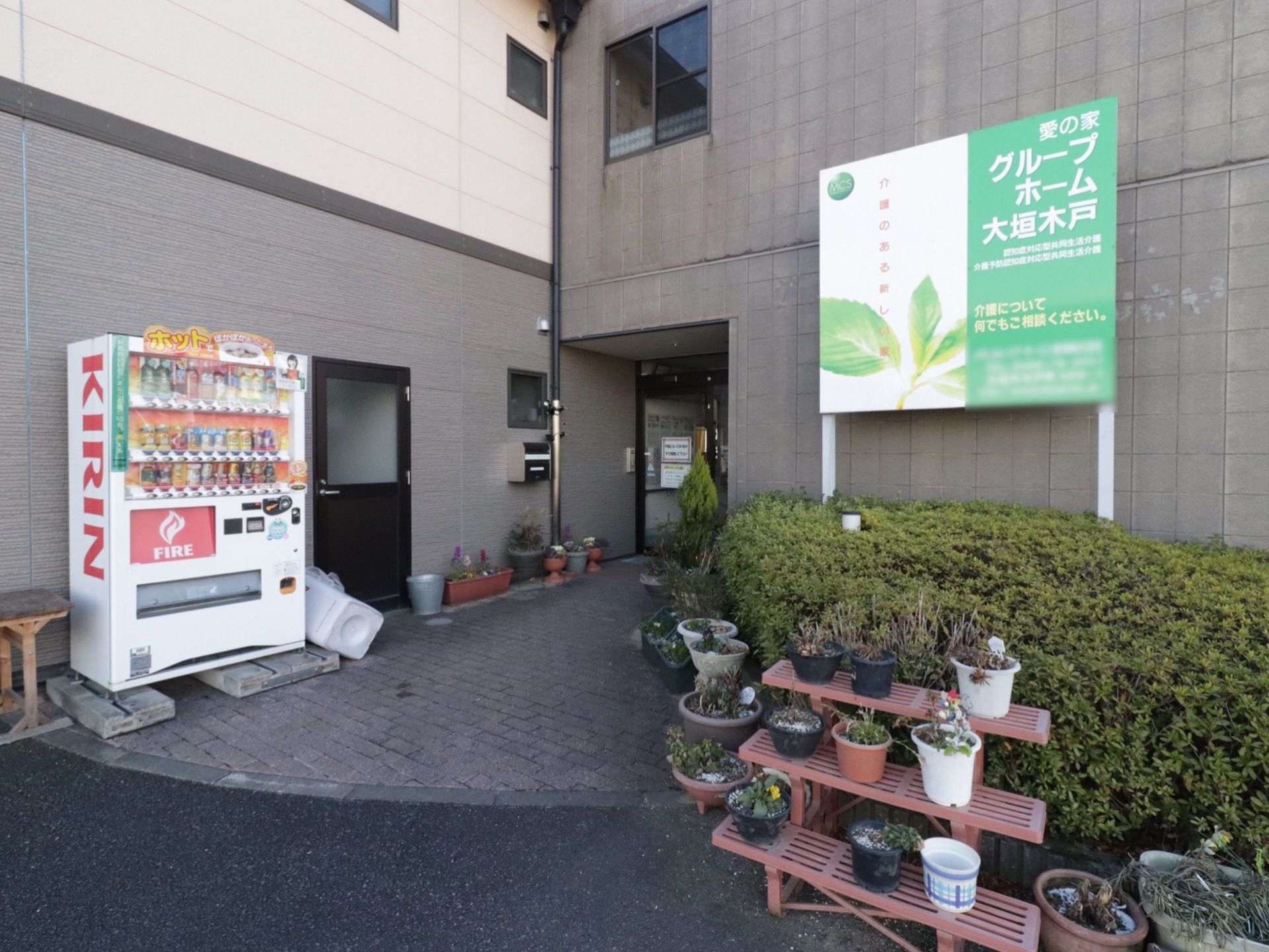 道路との境目までタイルが敷かれているエントランスである。自販機が設置されており、複数のプランターが置かれている。
