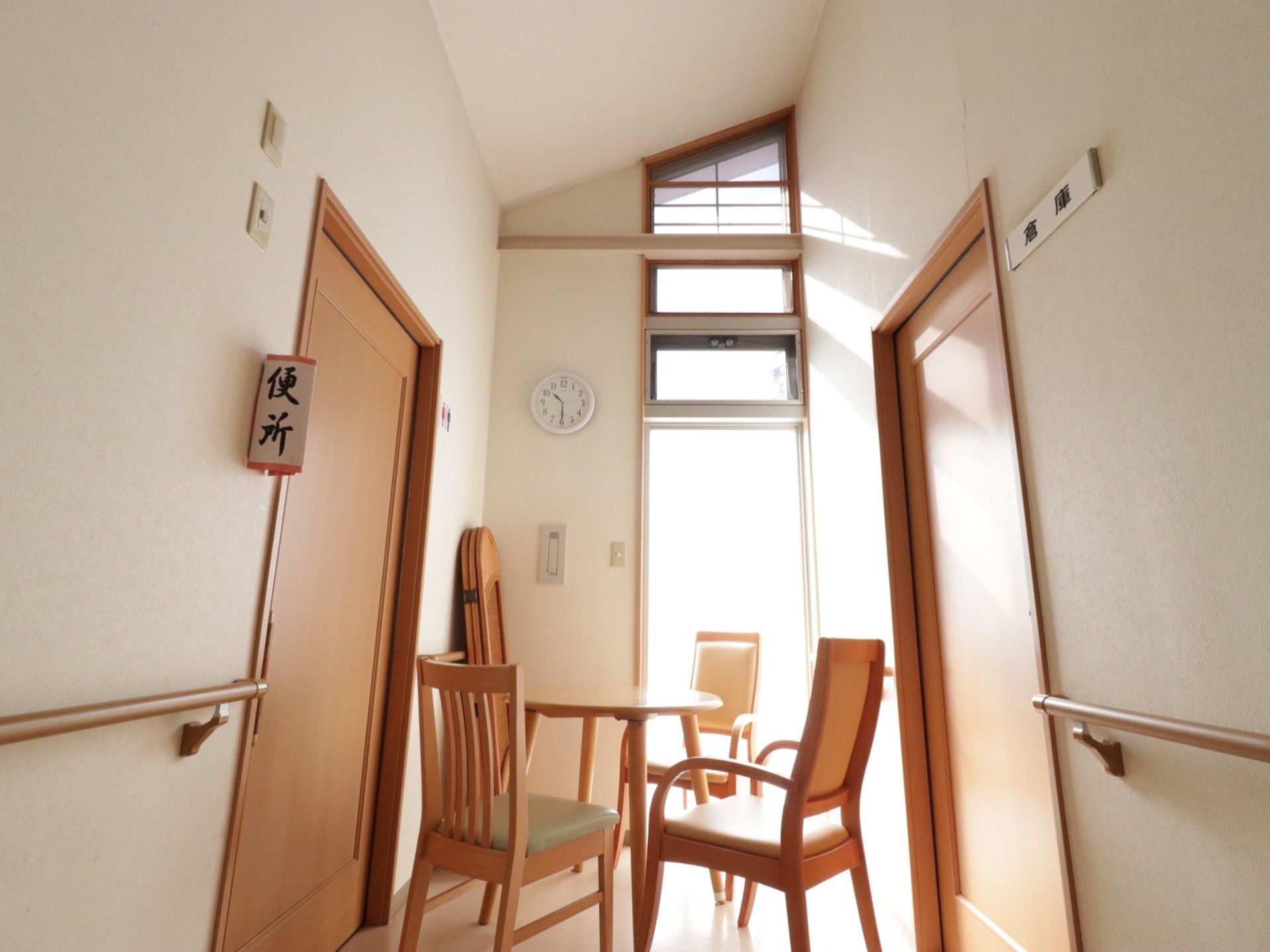 廊下の一角には、丸テーブルと３脚の椅子が置かれた談話スペースが設けられている。左にトイレ右に倉庫のドアがある。