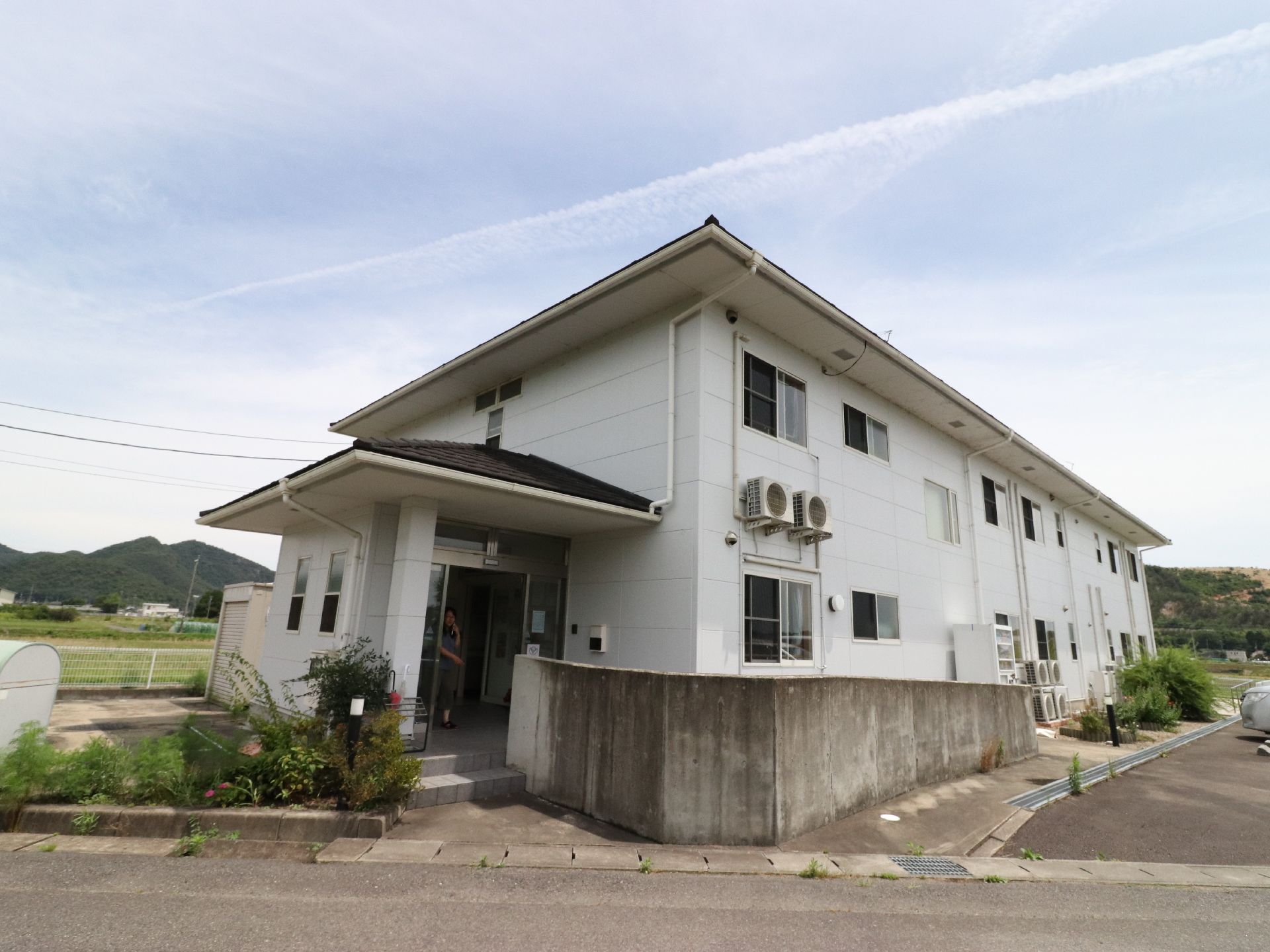 2階建ての白い建物。まわりは田畑に囲まれており、とてものどかな環境になっている。駐車スペースも広々としている。
