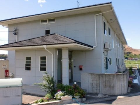グレーの外壁で切妻屋根が載った２階建ての建物は、玄関脇に小さな植え込みがあり、白い花が咲いている。塀の際にも花鉢が並んでいる。