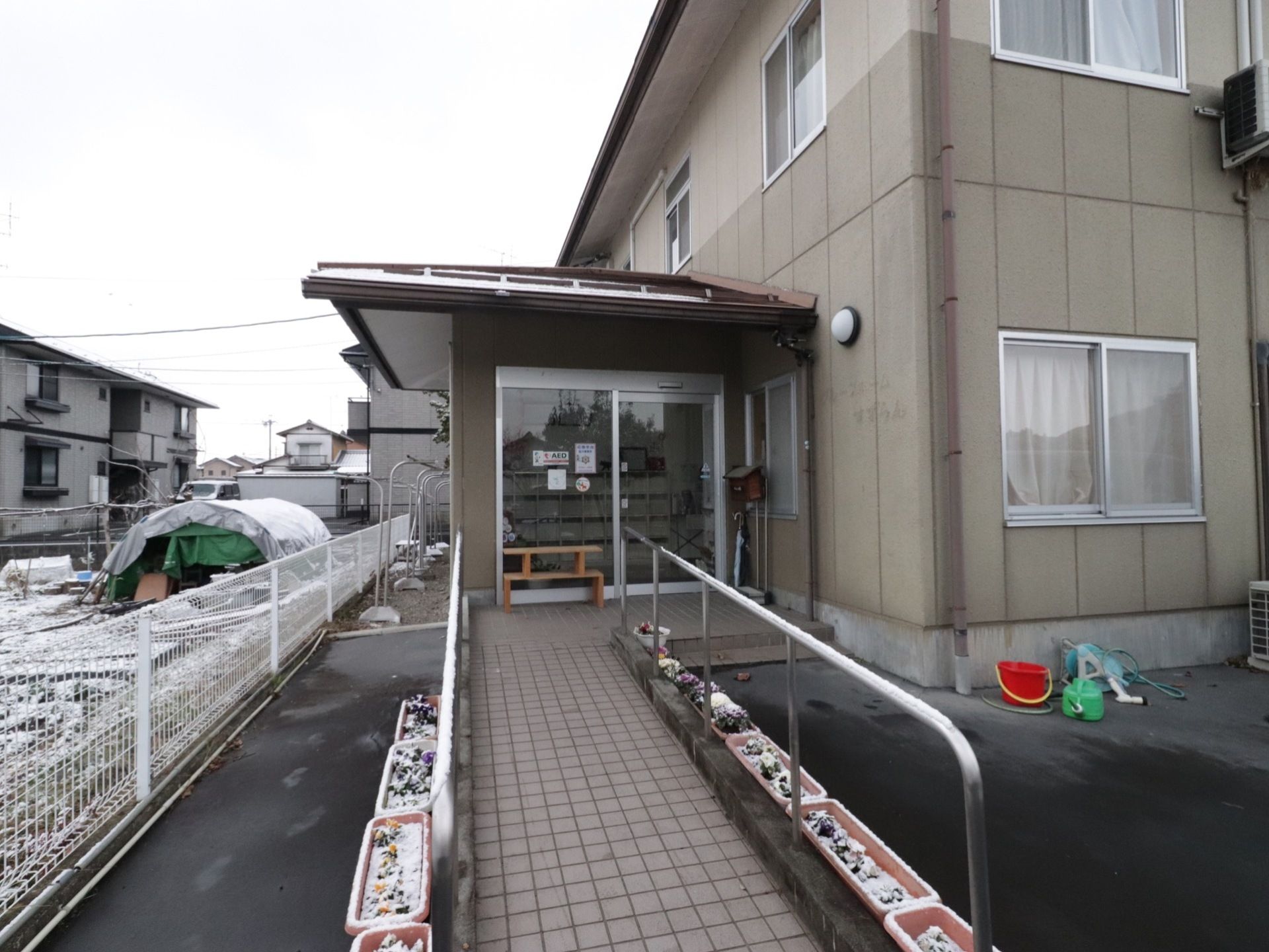 ２階建ての施設のエントランスには、手すり付きのスロープが設けられている。花が植えられたプランターが両サイドに並べてある。