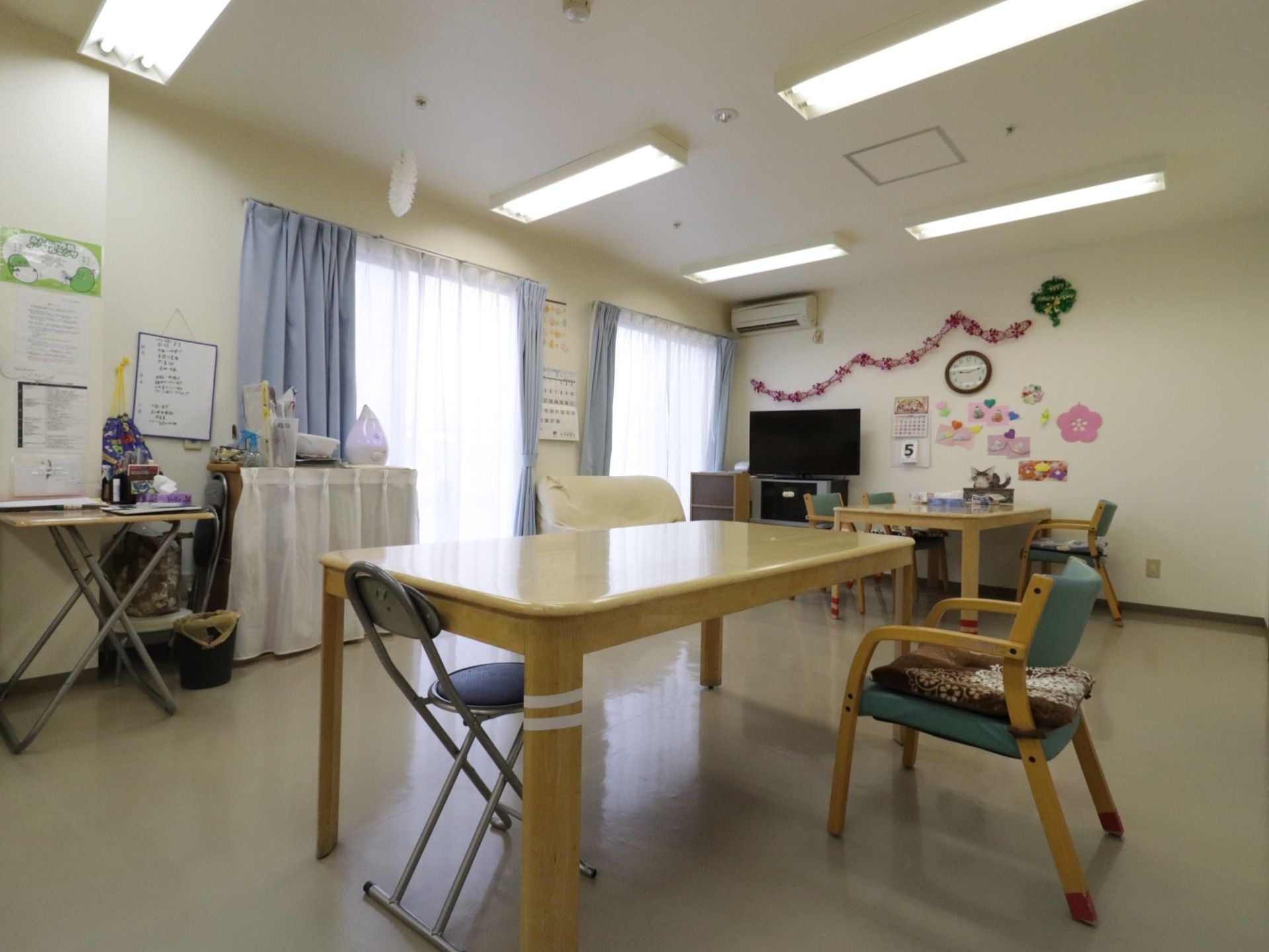 日中、窓から自然光が差し込む明るい食堂である。エアコンや照明が完備されており、ダイニングテーブルやテレビ、ソファが置かれている。