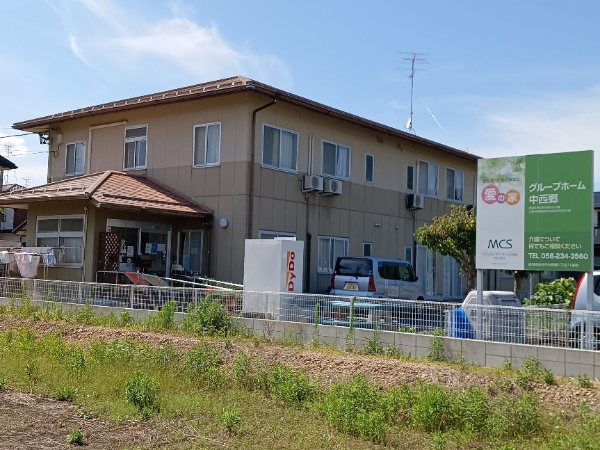２階建ての建物前に設けられた駐車場には、施設名が書かれた大きな看板がフェンス際に立っている。自販機が設置されている。
