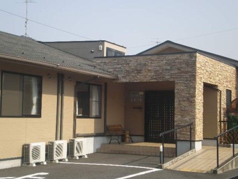 壁が茶色で屋根が黒い平屋の建物で、玄関部分がタイル貼りで柱があり、ひさしを支えている。手すり付きのスロープと階段が設けられている。