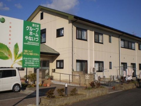 道路に面したベージュの外壁の２階建で、道路から玄関までは手すり付きのスロープをご利用いただける。玄関にはひさしが付いている。