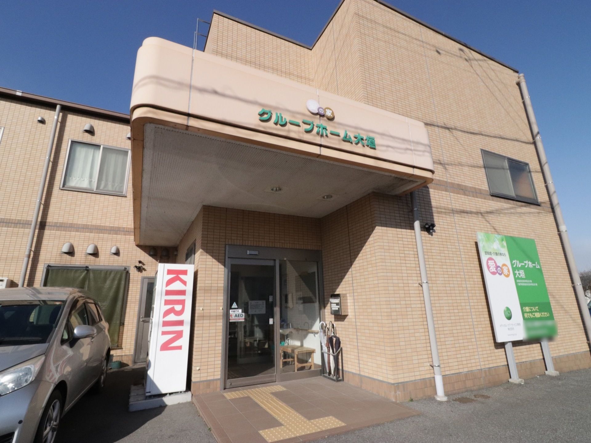 大きなひさしが付いたエントランスである。足元には、誘導ブロックが設置されている。傘立てが置かれている。