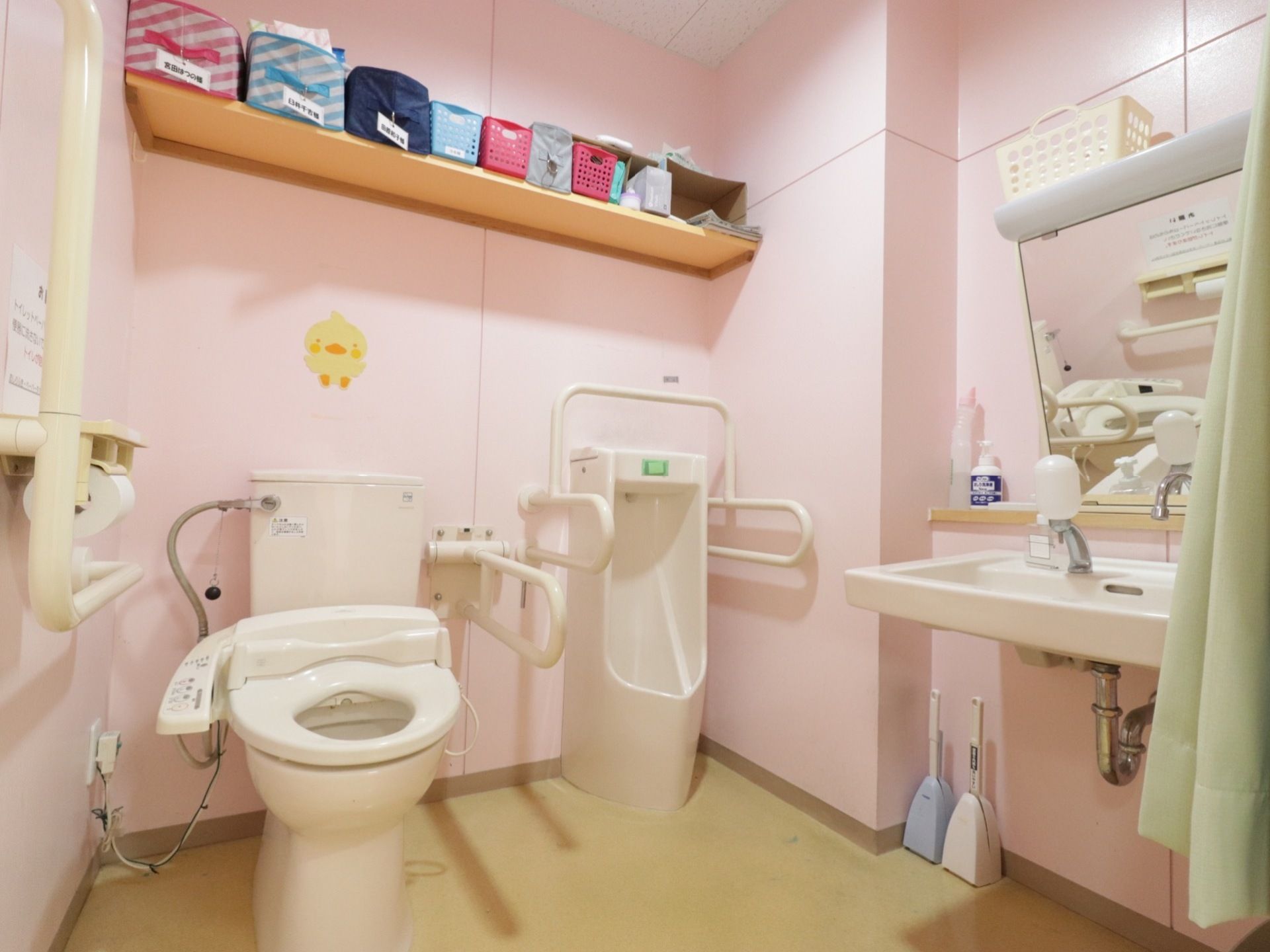 広々としたトイレには、洋式便器と男性用小便器が並んで設置されている。洗面台も備わっている。棚や手すりが壁に取り付けられている。