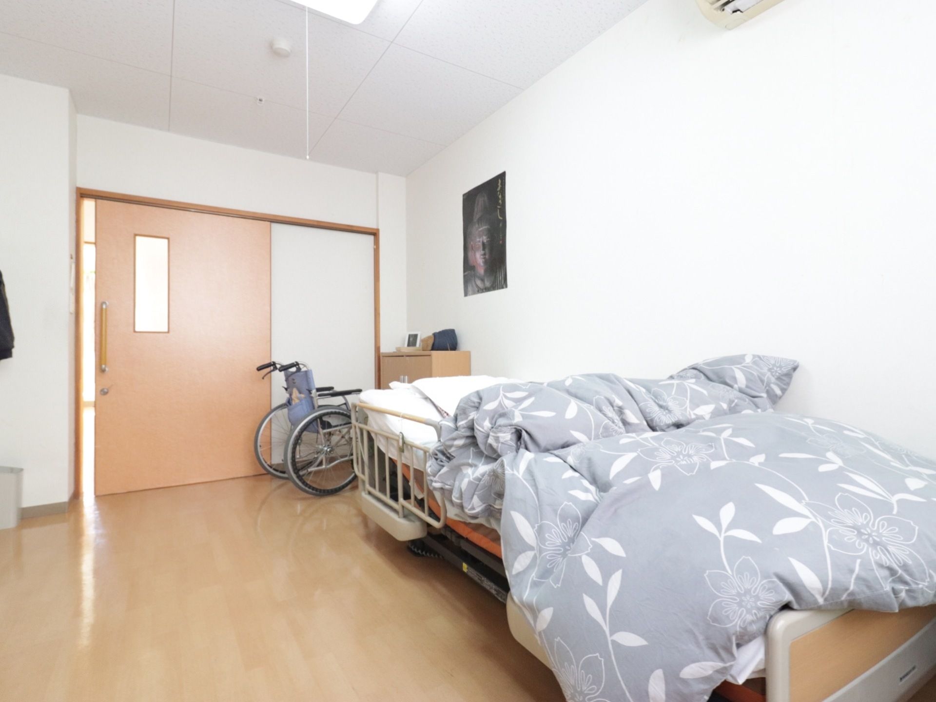 広い間取りの居室である。壁際に介護ベッドや車椅子、収納家具が置かれている。壁には、ポスターが貼られている。