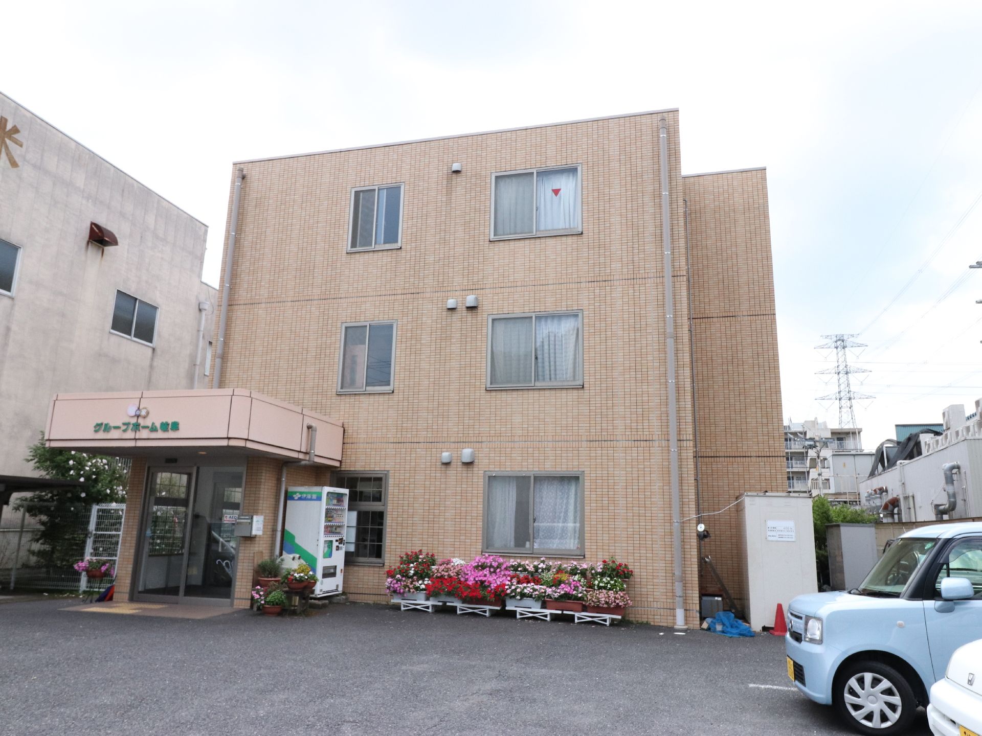 施設を正面から見た風景。3階建てで、1階左手が出入り口になっている。手前は駐車場のような空間で、建物の前にはプランターが並んでいる。