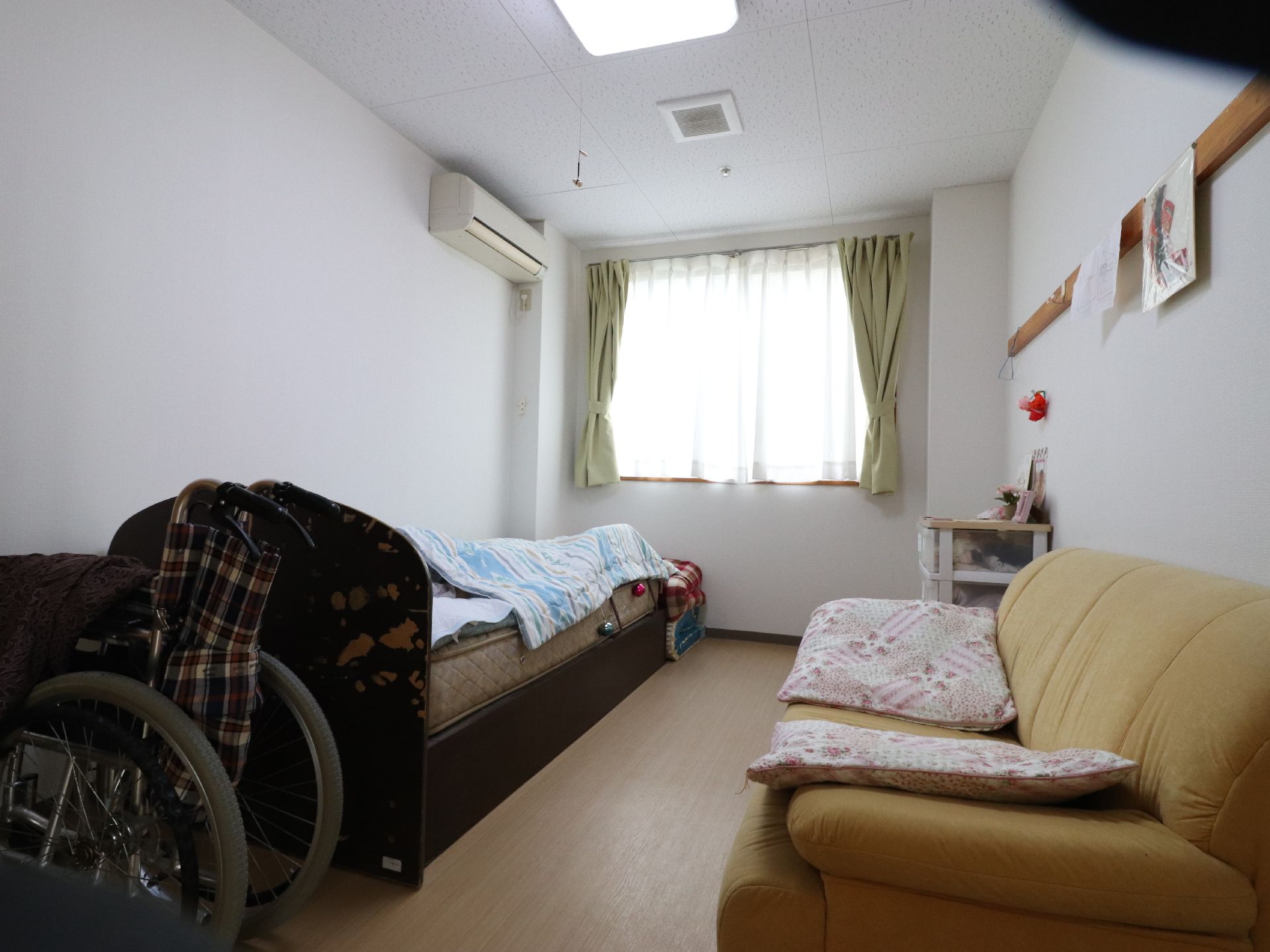 窓からの陽射しがふんだんに差し込む居住空間。右手にベッド、左にはソファーが置かれている。手前には車椅子が見受けられる。