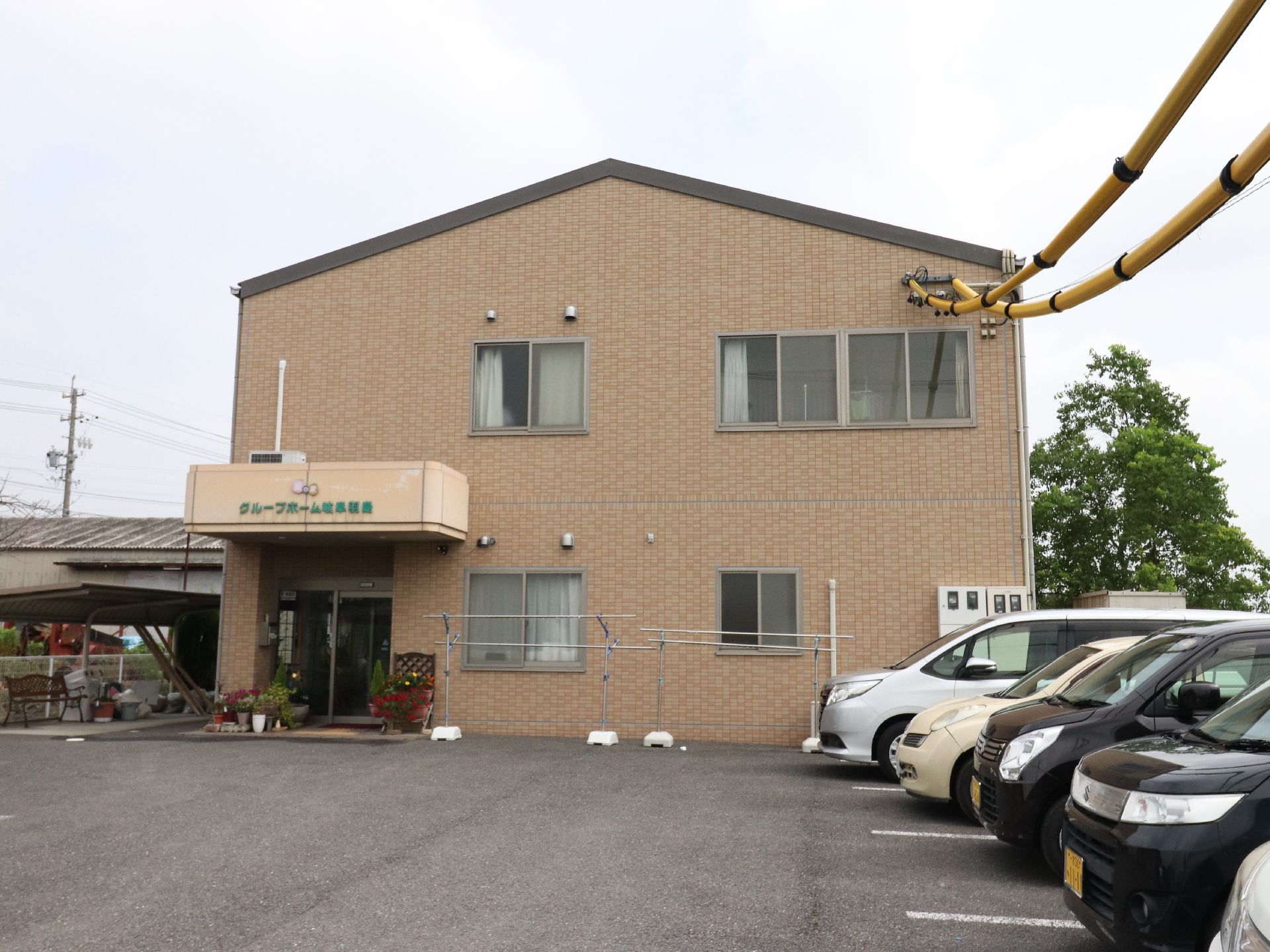 茶色の外壁が落ち着きを感じさせる建物の全景。三角屋根の2階建てで、手前のスペースは駐車場になっている。