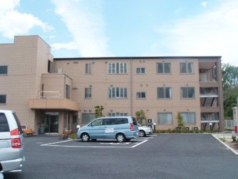 ベージュの外壁の３階建ての建物は、左側にひさし付の玄関があり、右手には非常階段がある。中央には駐車スペースがある。
