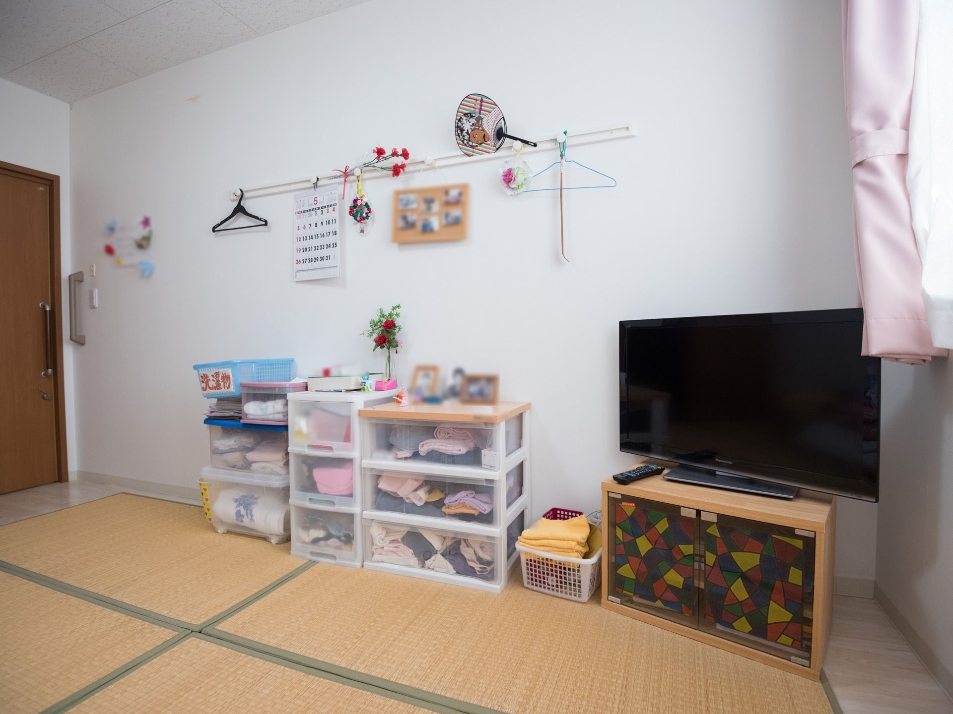 畳が敷かれている和室である。テレビ台の扉はカラフルなステンドガラス風になっており、壁には写真やカレンダーが掛けられている。