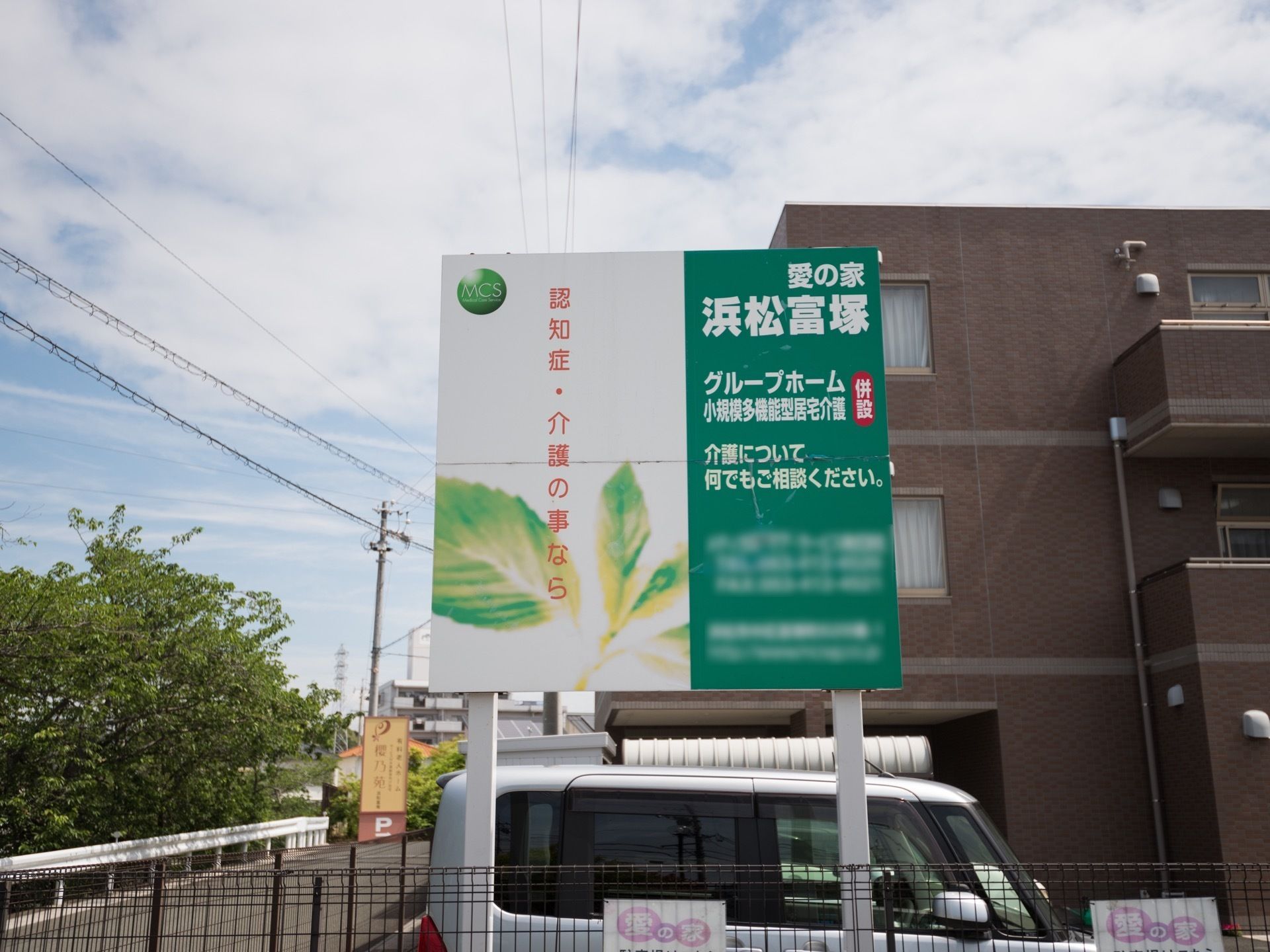 グループホームの看板が立てられている。奥には３階建ての茶色い建物があり、その前に車が駐車されている。
