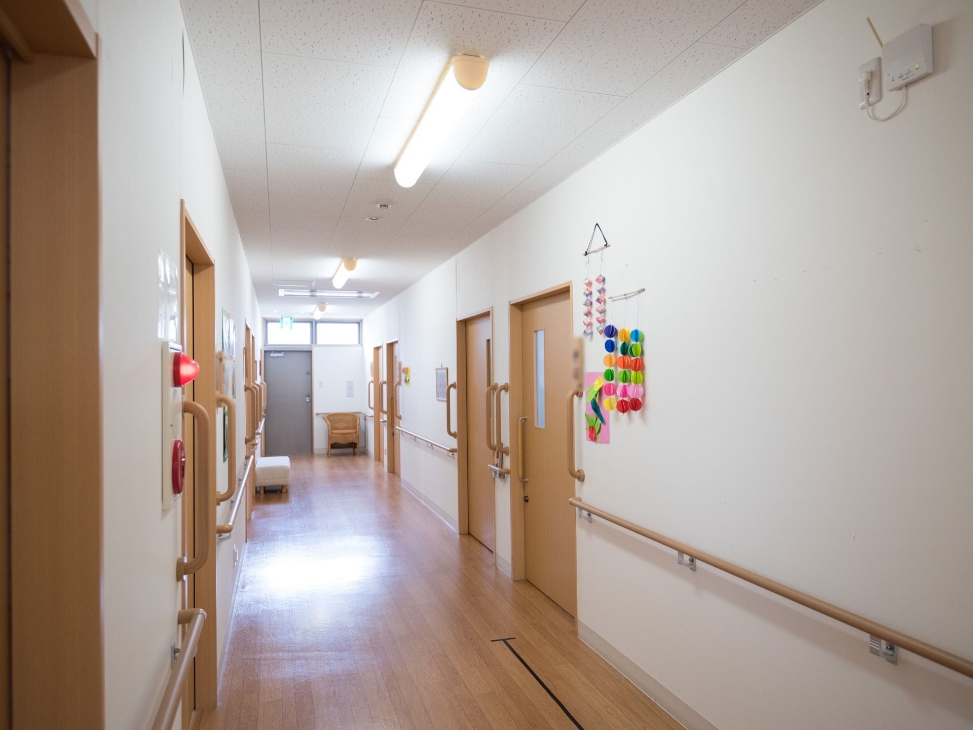 白い壁に木製の手すりが取り付けられている廊下である。両側には居室の入口が並んでいる。また、白色の長椅子が１脚置かれている。