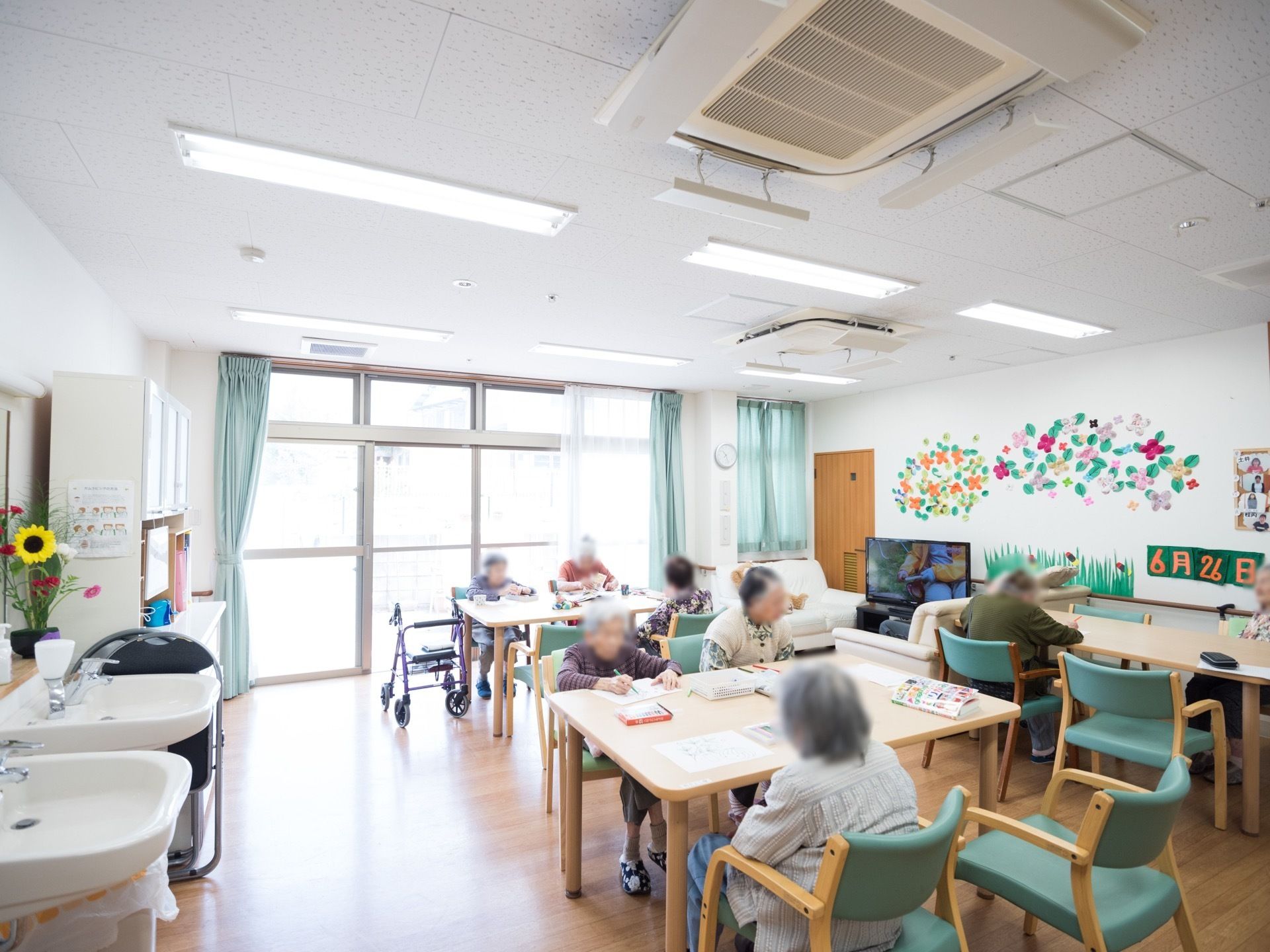 複数のテーブルが設置され、そこに入居者様が座り作業をされている様子である。壁には複数の飾りがされている。