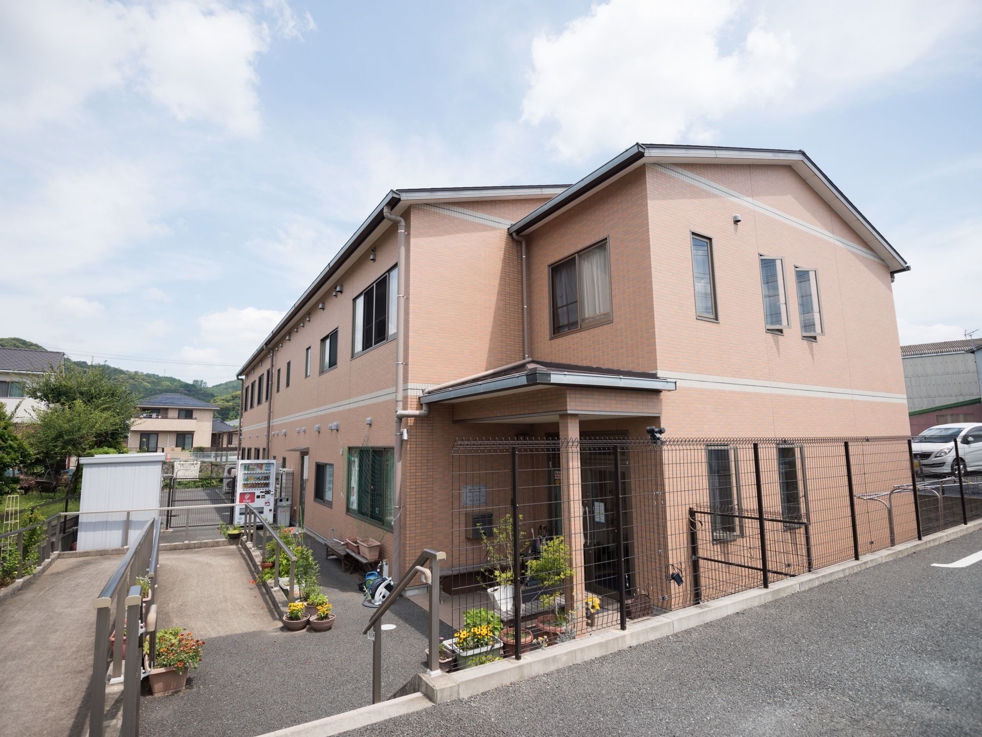 ２階建ての建物で、窓や換気口が並んでいる。建物付近には自動販売機が設置されているほか、植物の鉢が複数並べられている。