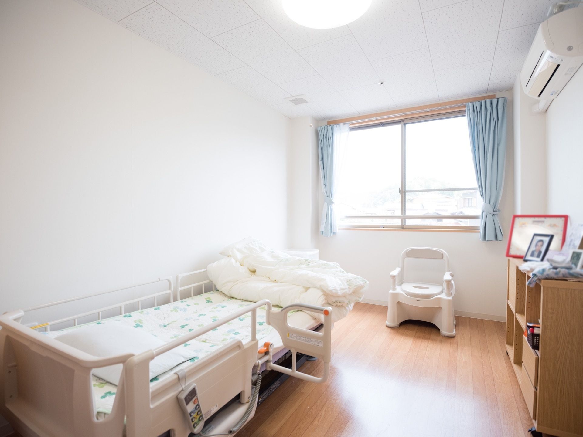 白を基調とした清潔感のある居室である。電動リクライニング式のベッドが１台壁側に置かれ、窓際には白い椅子が１脚ある。