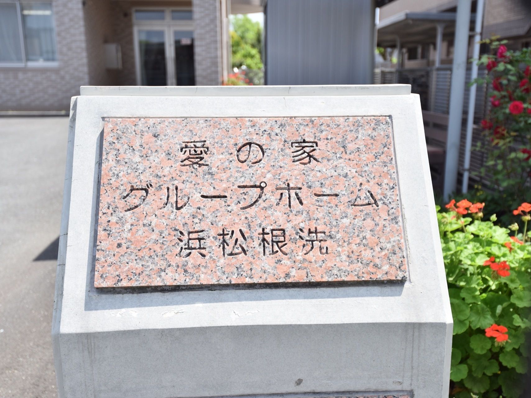 施設敷地内に表札が設置されており、後ろにある花壇に赤い花が咲いているのが見える。玄関横の外壁には、ポストが設置されている。