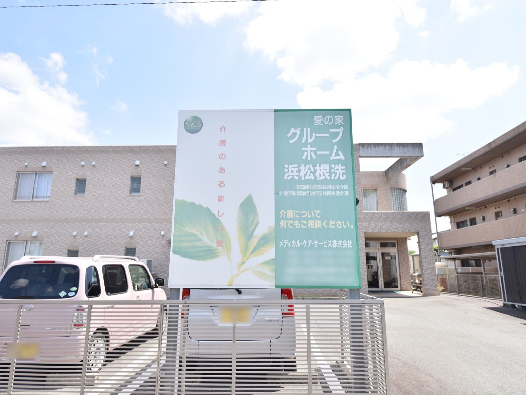 駐車場のフェンス上部に、看板が設置されている。施設はブラウンを基調とした、２階建ての建物となっている。