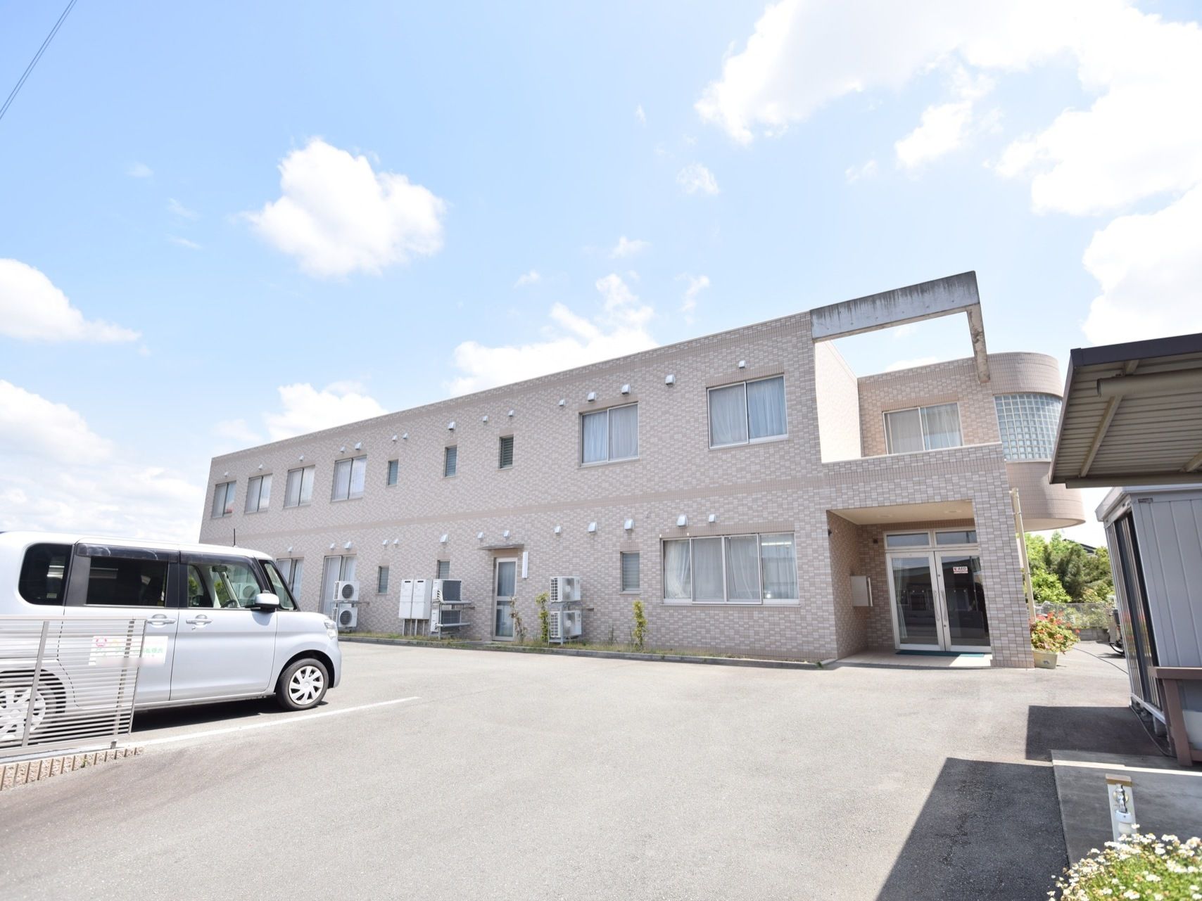 日当たりの良いホーム。建物外壁に沿って複数のエアコン室外機が置かれており、駐車場横には物置が設置されている。