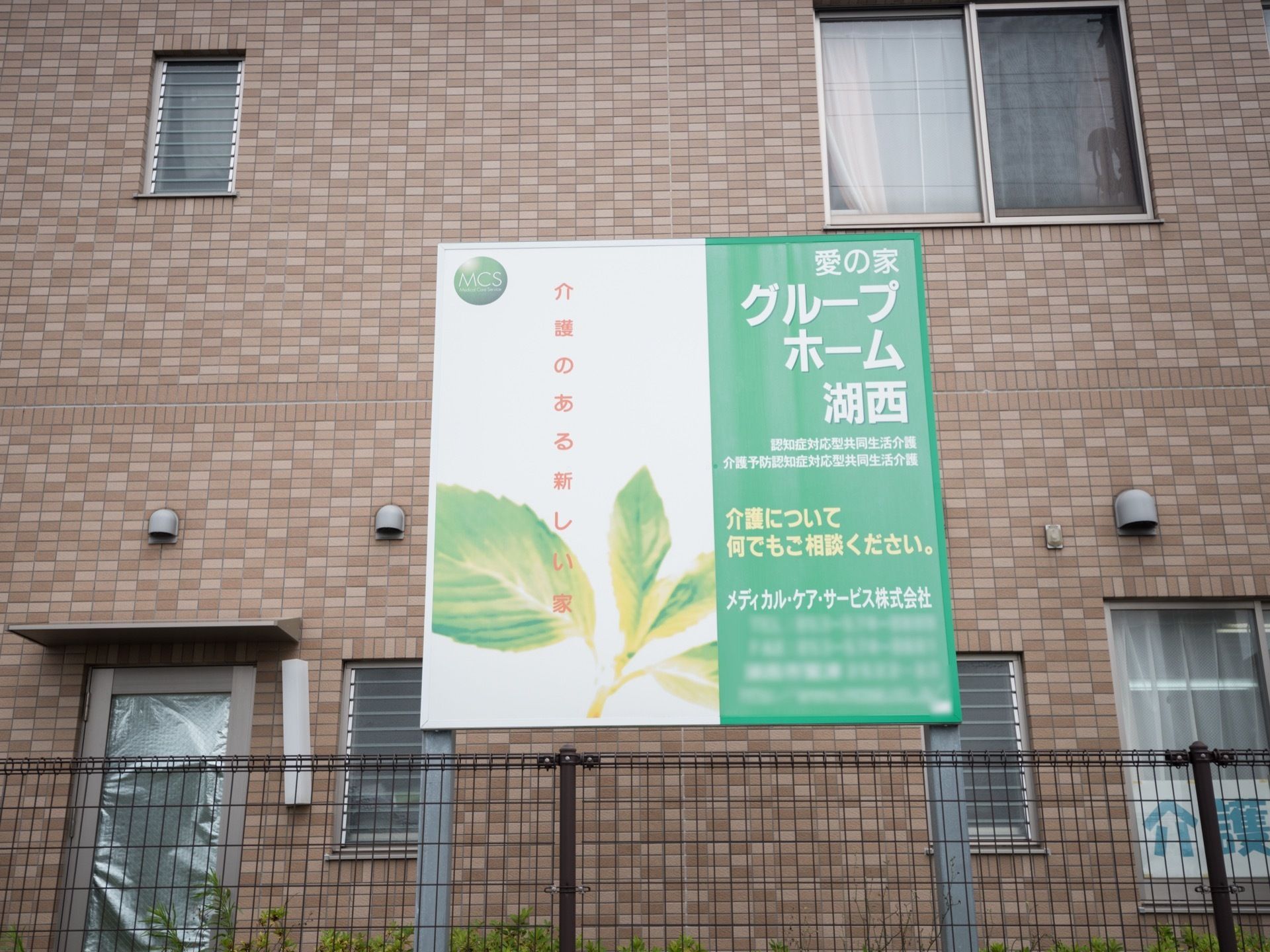敷地のまわりに、ダーク調のフェンスが設置されている。白と緑の施設看板も設けられている。外壁は茶系で落ち着いた色合い。
