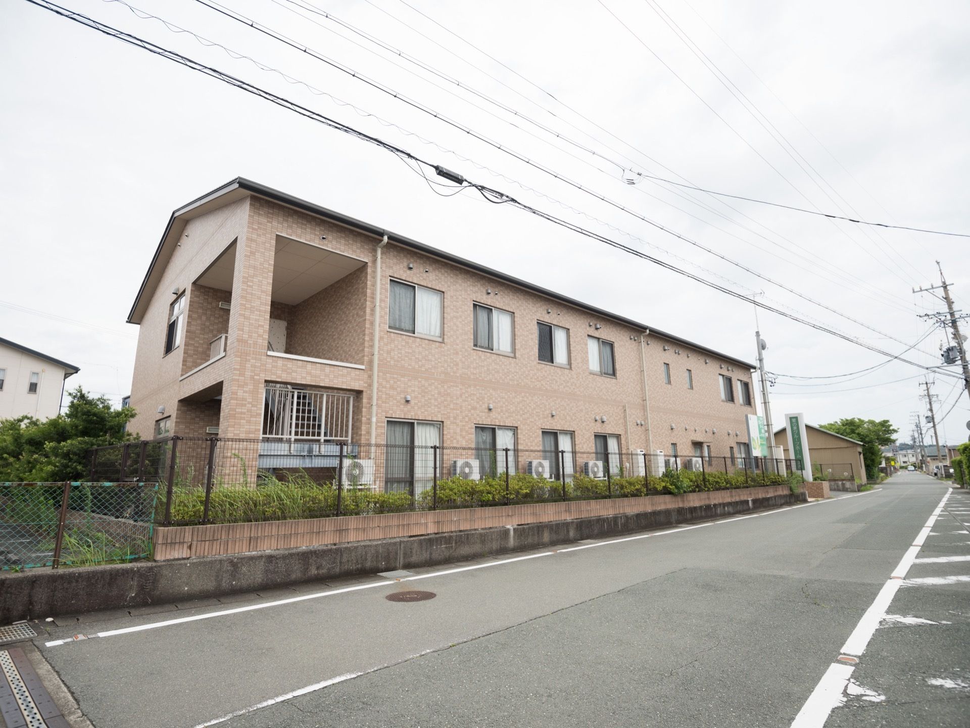 道路に面したところに立地する、２階建ての施設である。落ち着いたブラウンの外壁。まわりにフェンスが立っている。