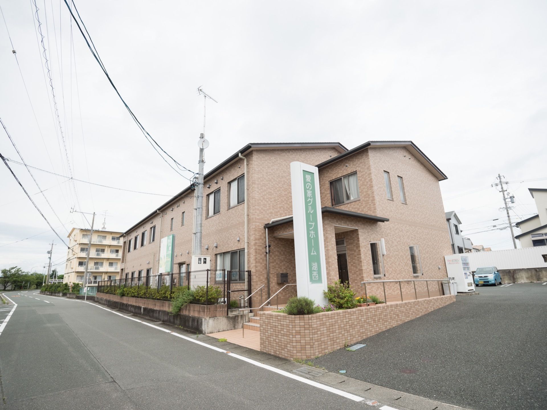 茶色のレンガ調に施された、落ち着いた色合いの外壁である。フェンス前に緑が設けられている。目の前には、車道が走っている。