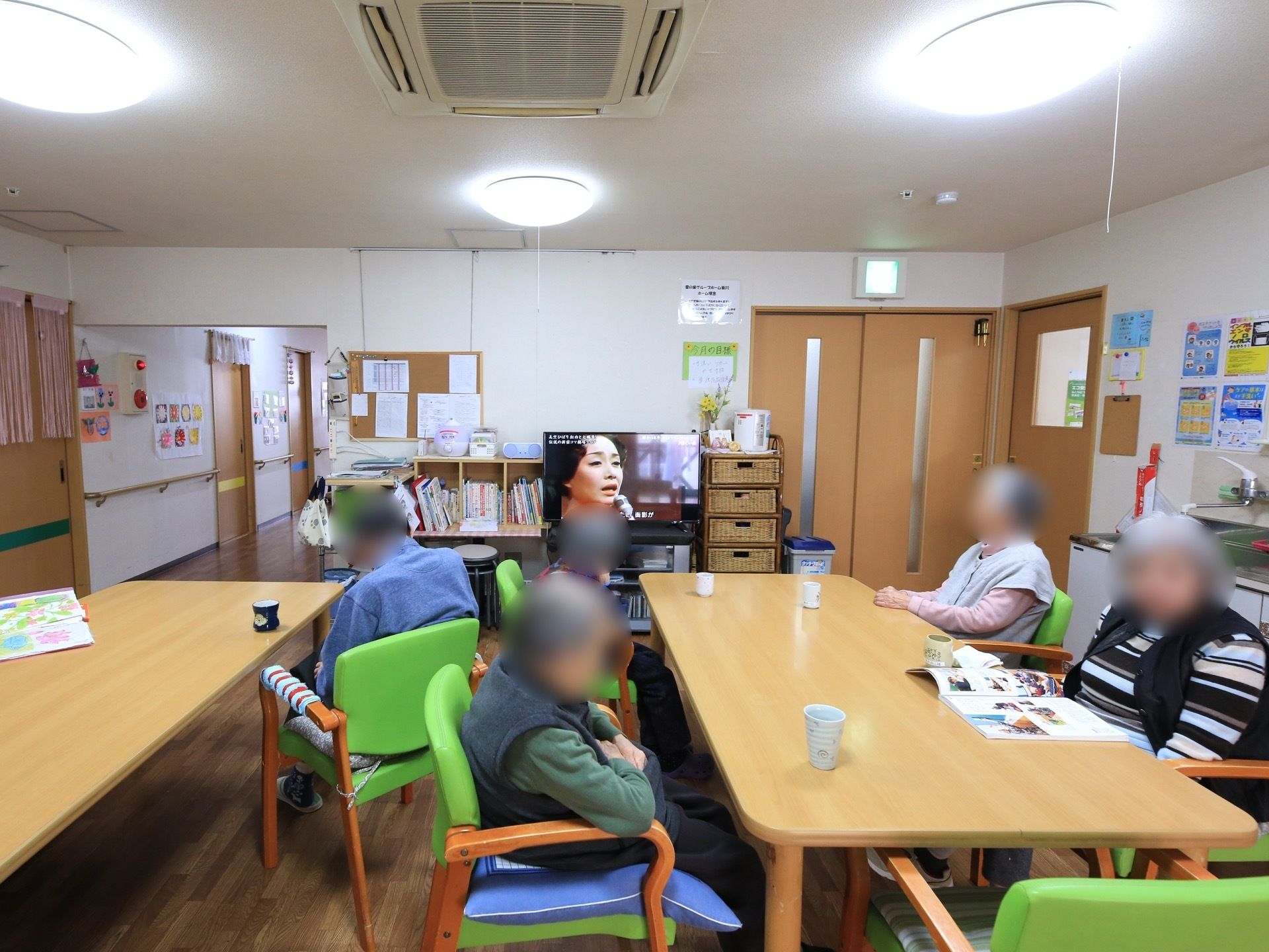 木製のテーブルセットが設置されている。黄緑の椅子が並べられ、入居者様がくつろいでいる。奥にテレビが置かれている。