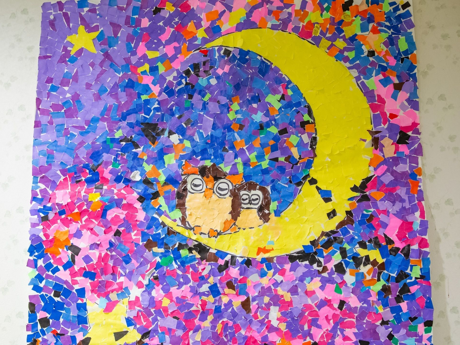 色鮮やかな紙を使って作られた大きなちぎり絵が壁に飾られている。月や星、２羽のフクロウが描かれている。