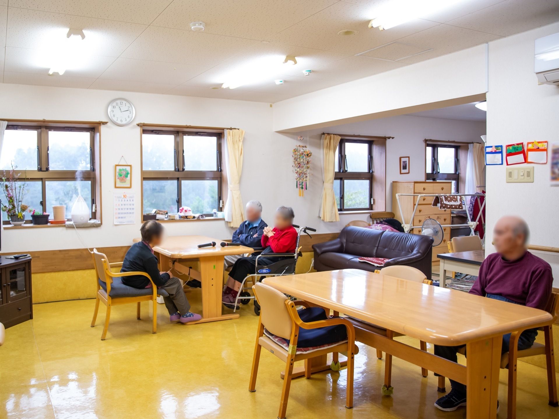 広い部屋に、ダイニングテーブルが２セットあり、入居者様が腰かけている。ソファや書き物デスクなども置かれている。