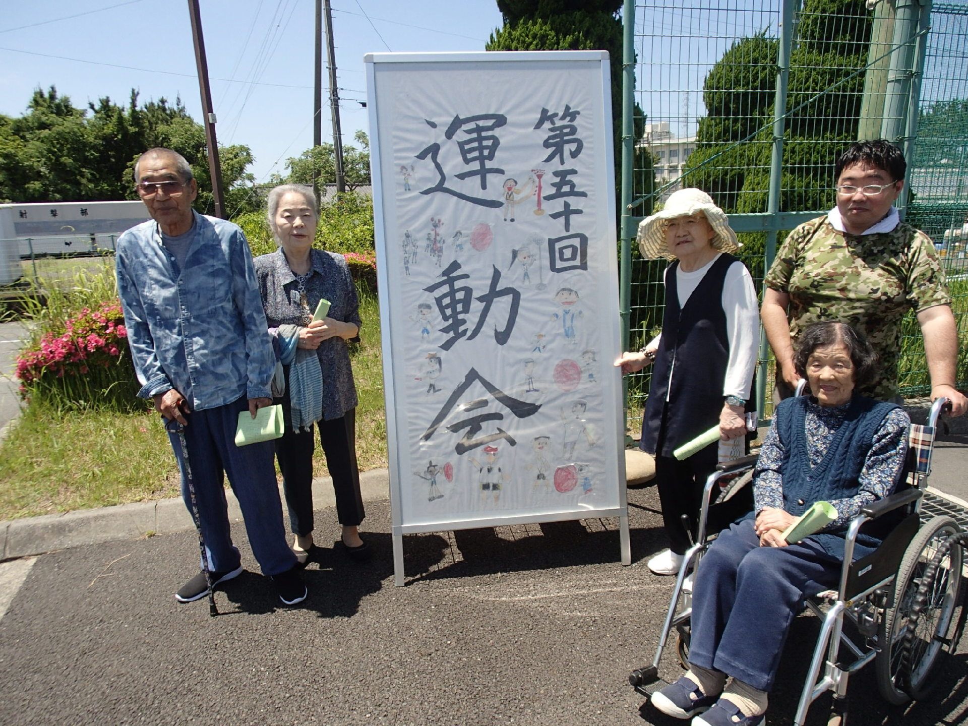 『運動会』の看板を囲むように高齢者が並んでいる。後ろでピンク色の花が咲いている