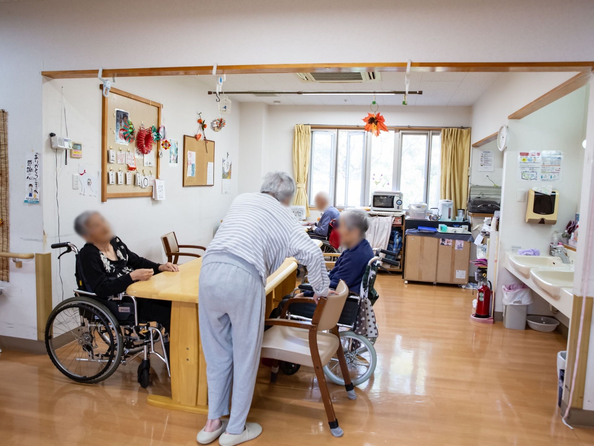 空調のついた部屋で、入居者様がテーブルについている。洗面台が２か所あり、奥にはポットや電子レンジが置かれている。