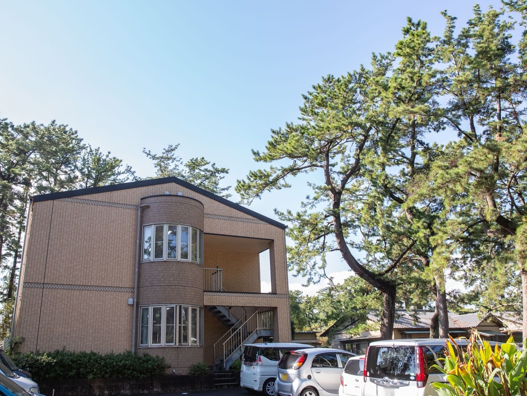 レンガ調の２階建ての建物がある。真ん中は円柱状にせり出している。建物は周りを背の高い木々に囲まれている。
