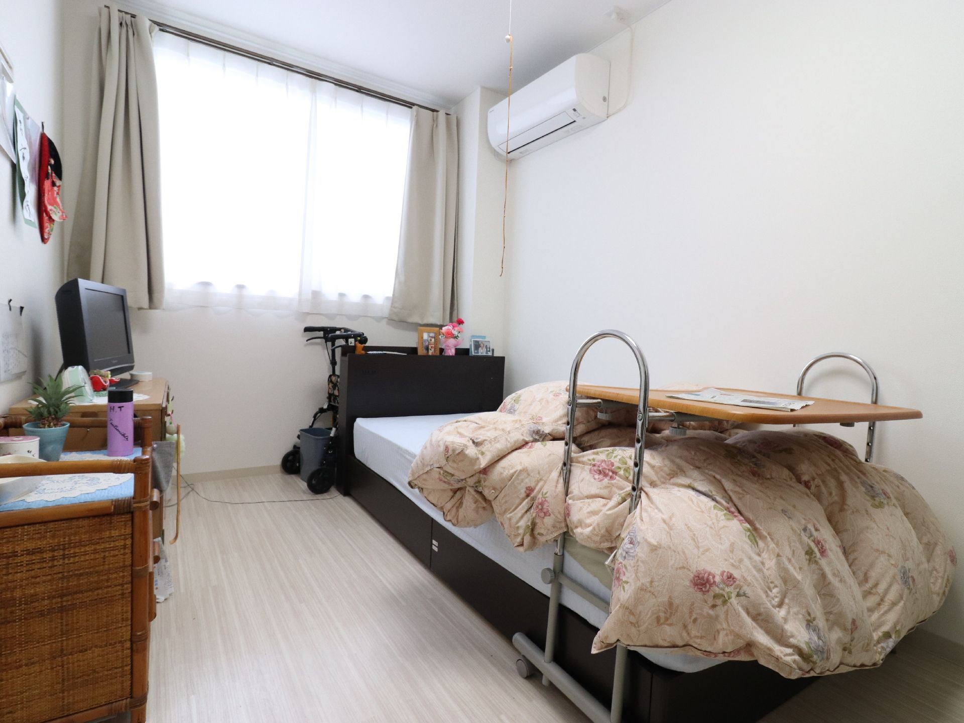 部屋に介護ベッドではない一般的なベッドが置かれている。籐の収納家具やテレビが並び、一般家庭のような居室。