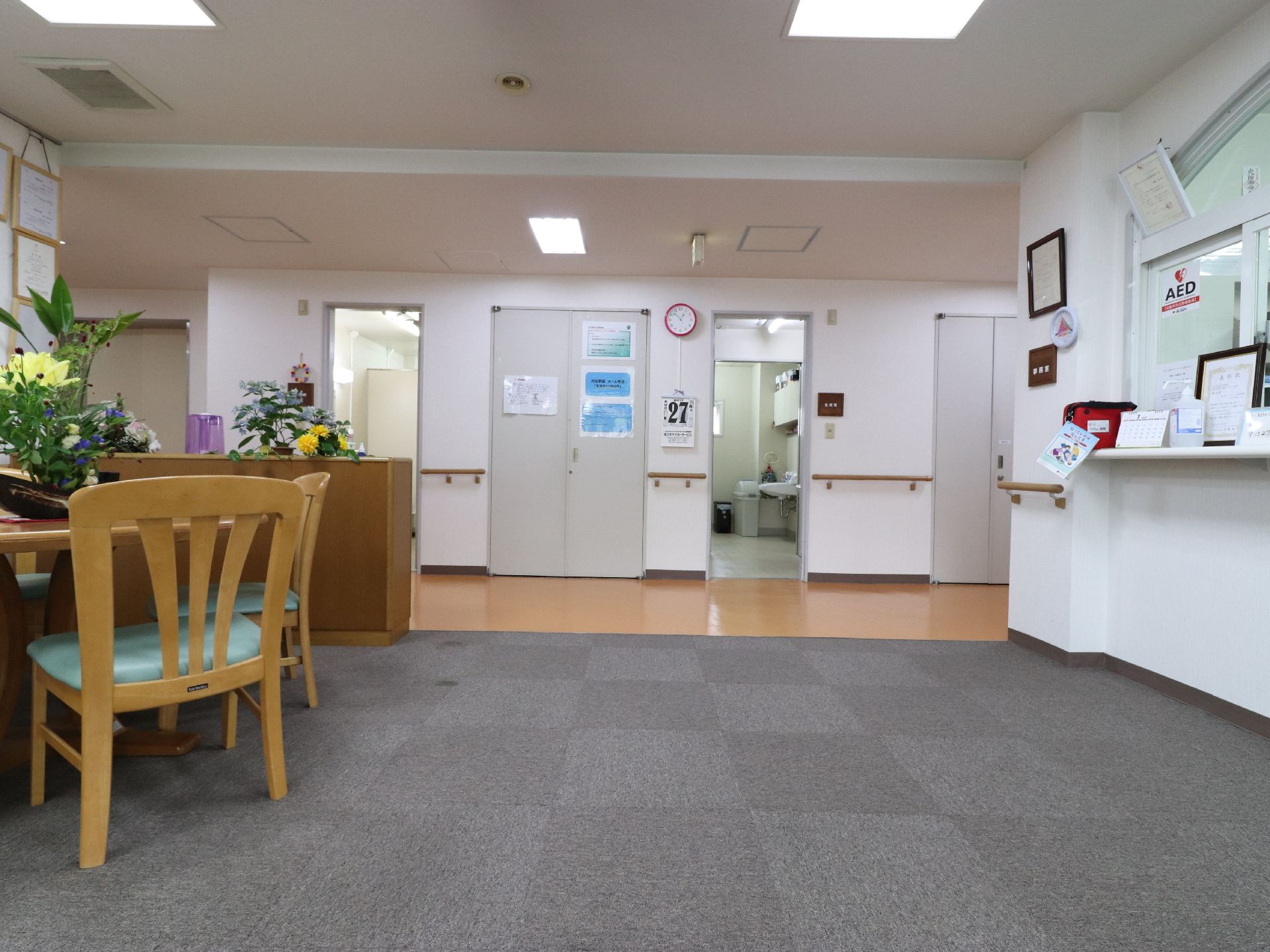 向かって右手に受付、正面に時計、左手に椅子のあるロビー。床はバリアフリーで、グレーのカーペットが敷かれている。