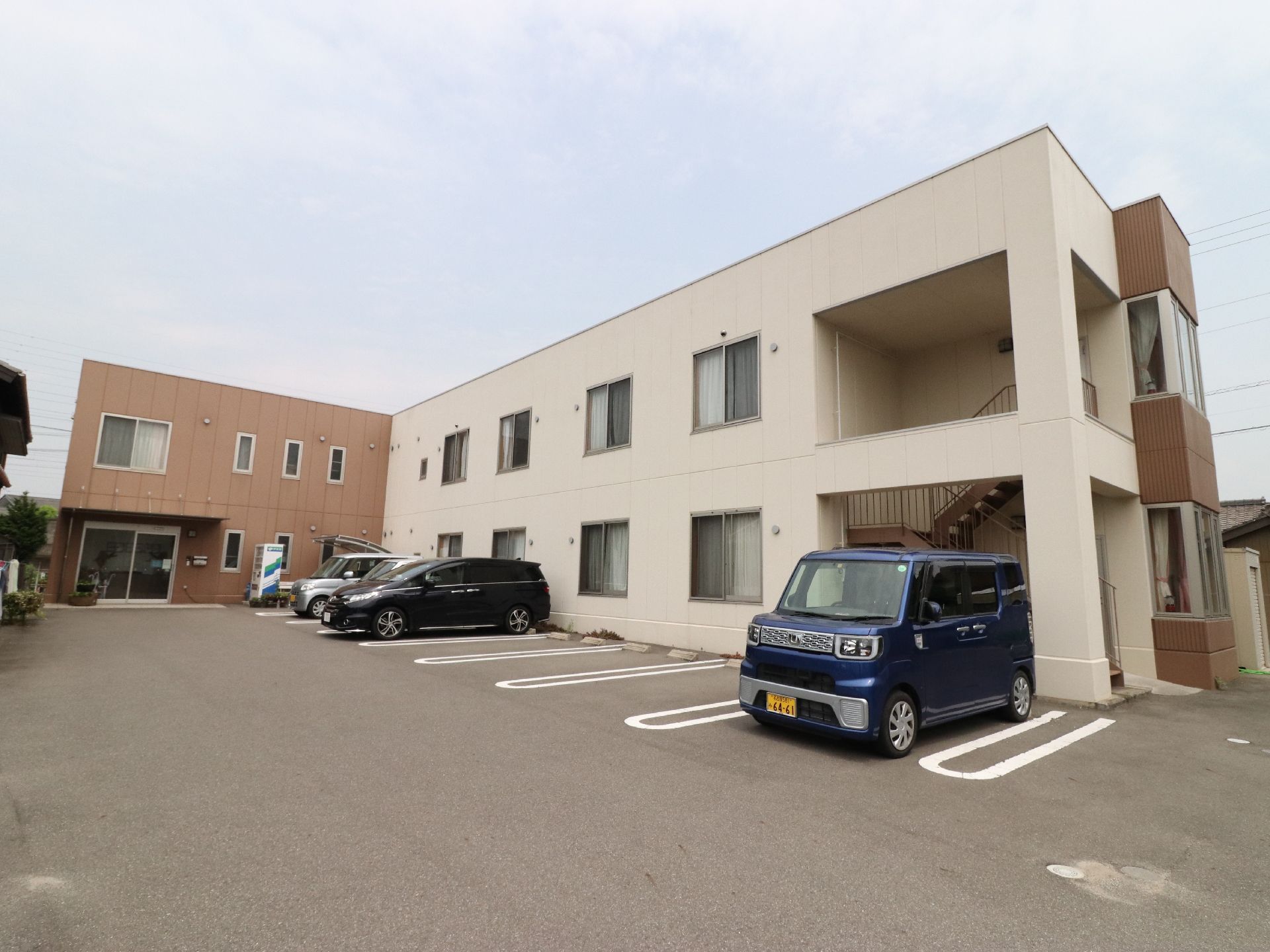 施設の建物は2階建てで、L字型の外構。棟によって壁の色がツートンカラーに分かれている。手前のスペースは駐車場になっている。