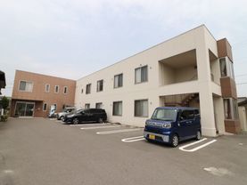 施設の建物は2階建てで、L字型の外構。棟によって壁の色がツートンカラーに分かれている。手前のスペースは駐車場になっている。