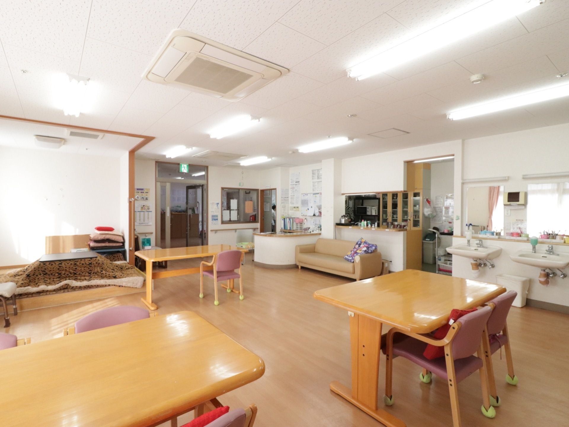 室内にはテーブルとイスが大きく間隔をとって配置されている。写真中央の壁際にはソファが置かれている。その奥には厨房がみえる。