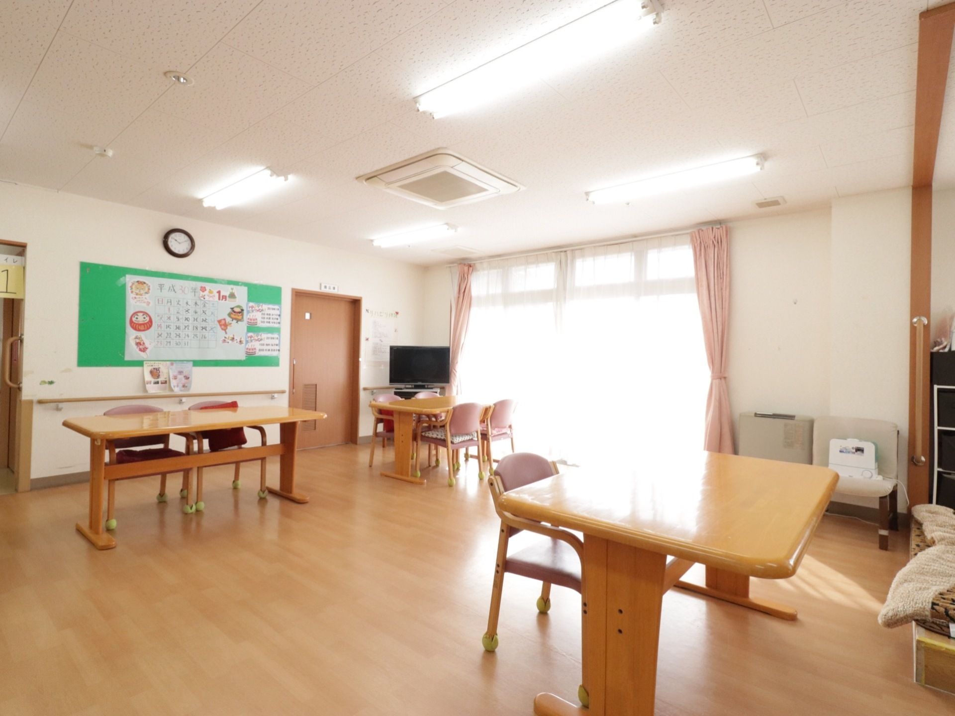 室内には食卓が置かれている。奥の壁にはカーテンのかかった掃き出し窓が設置されている。その付近には薄型のテレビがある。
