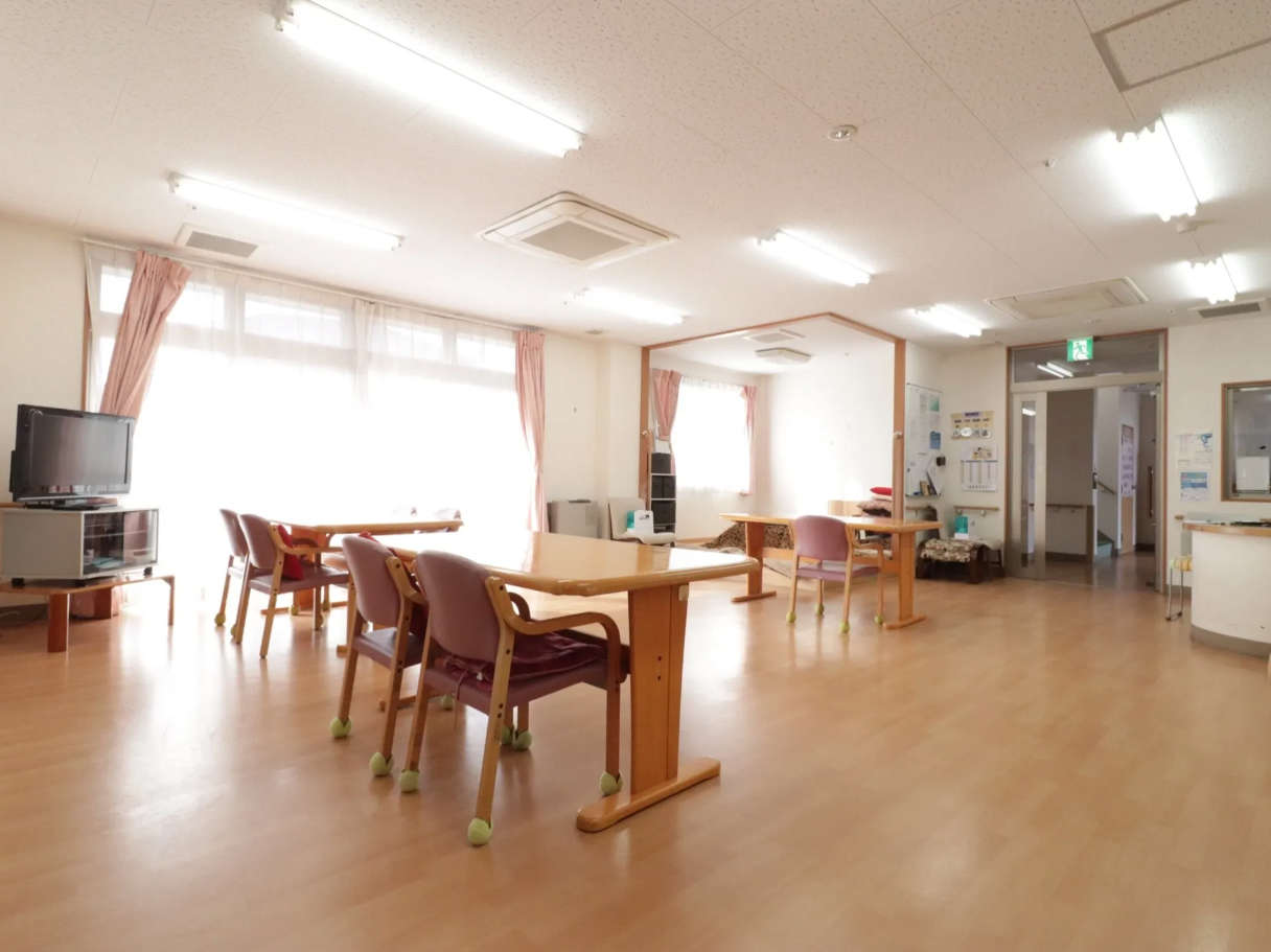 画像は食堂の一角の畳スペースである。中央にコタツ式テーブルが置かれている。壁にはカーテンの掛けられた長方形の窓が設置されている。