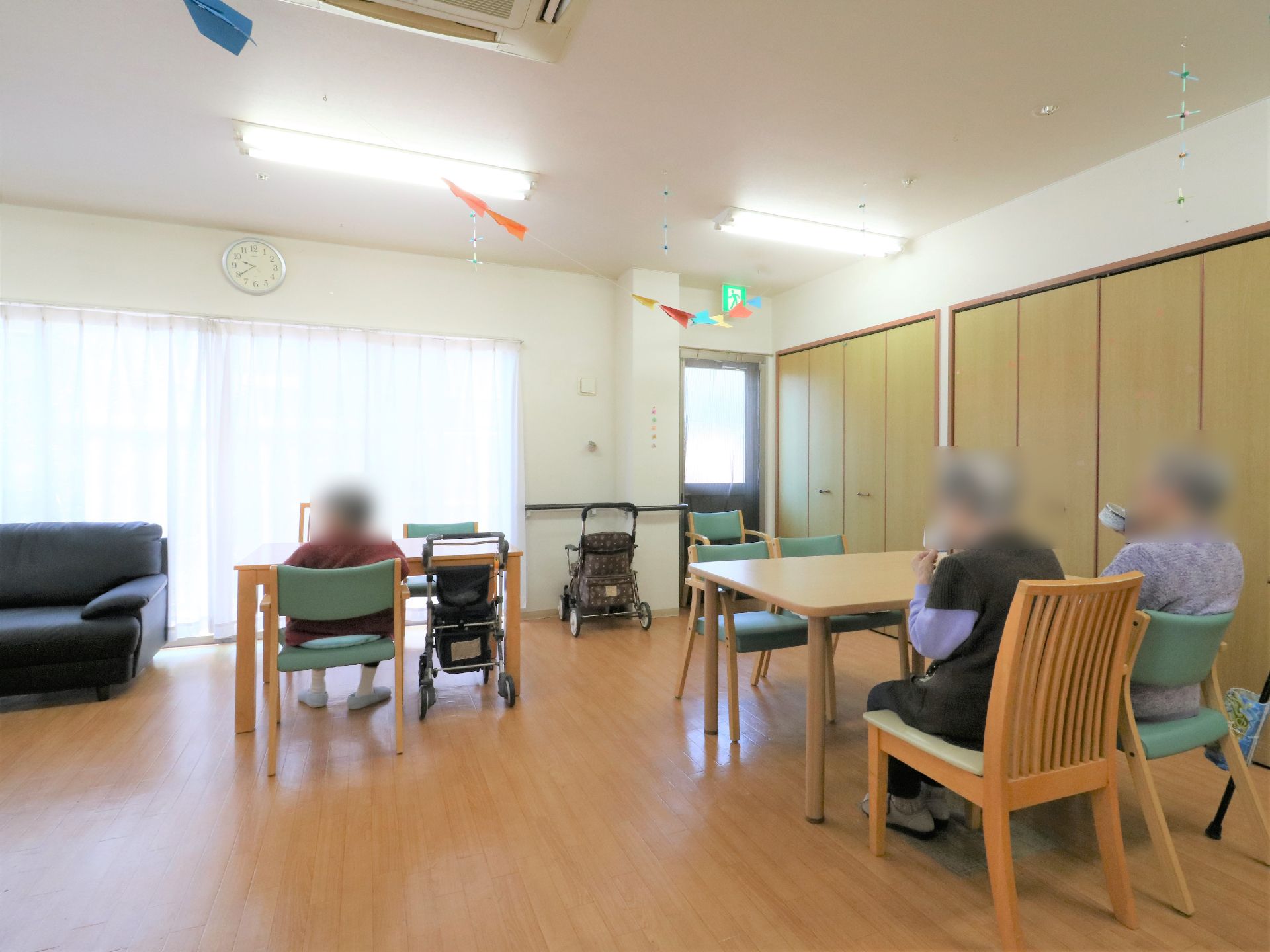 広いリビングにはテーブルと椅子が置かれ3人に入居者が座っている。壁には大きなクローゼットがあり窓際にはソファーも置かれている。