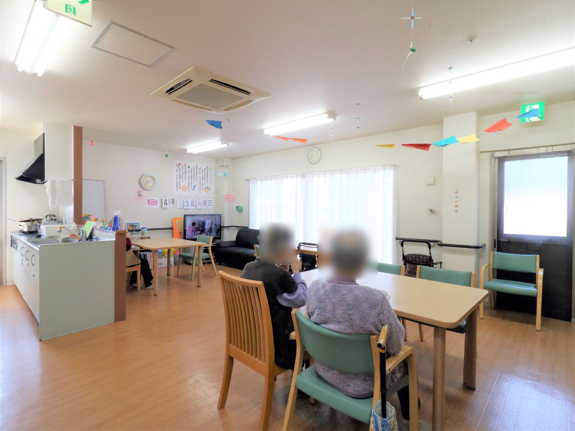 リビングの端にはテレビが設置されていて壁には日付が判りやすく貼り紙されている。手前にはキッチン設備などもあるのが見える。
