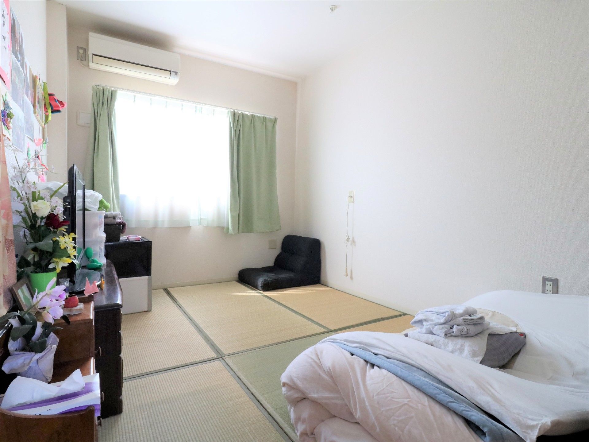 居室は洋室タイプのワンルームに床は畳が敷かれている。奥には窓があり座椅子や棚、箪笥などが置かれ手前には布団が畳まれている。